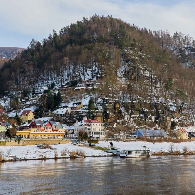 Schneebedeckte Hügel mit Bäumen, darunter ein Dorf am Wasser mit Booten.