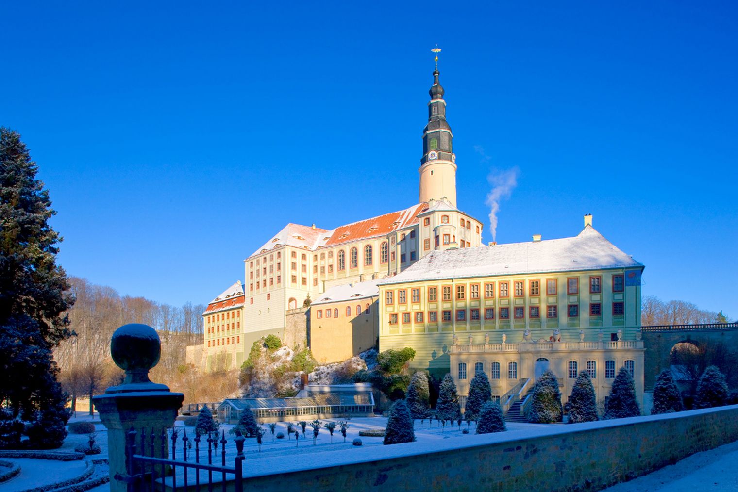 Schneebedecktes Schloss auf einem Hügel, umgeben von Bäumen und klarem blauen Himmel.