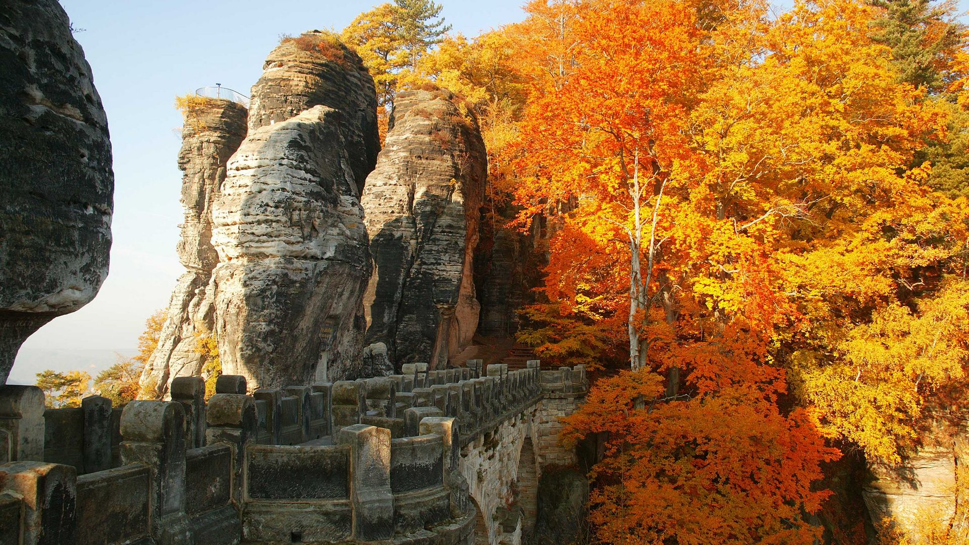 Herbstliche Basteibrücke, umgeben von Felsen und buntem Laub unter bewölktem Himmel.
