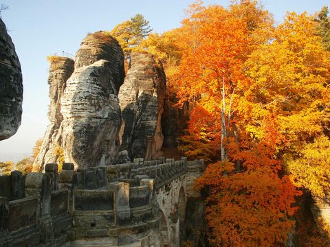 Herbstliche Basteibrücke, umgeben von Felsen und buntem Laub unter bewölktem Himmel.
