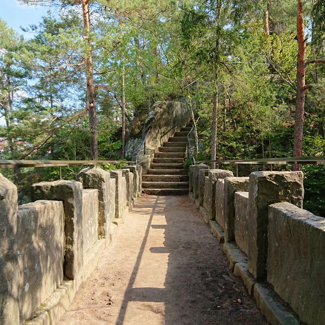 Teufelsbrücke am Hockstein mit Steinmauer, umgeben von Bäumen und Sträuchern, führt zu einer Treppe.