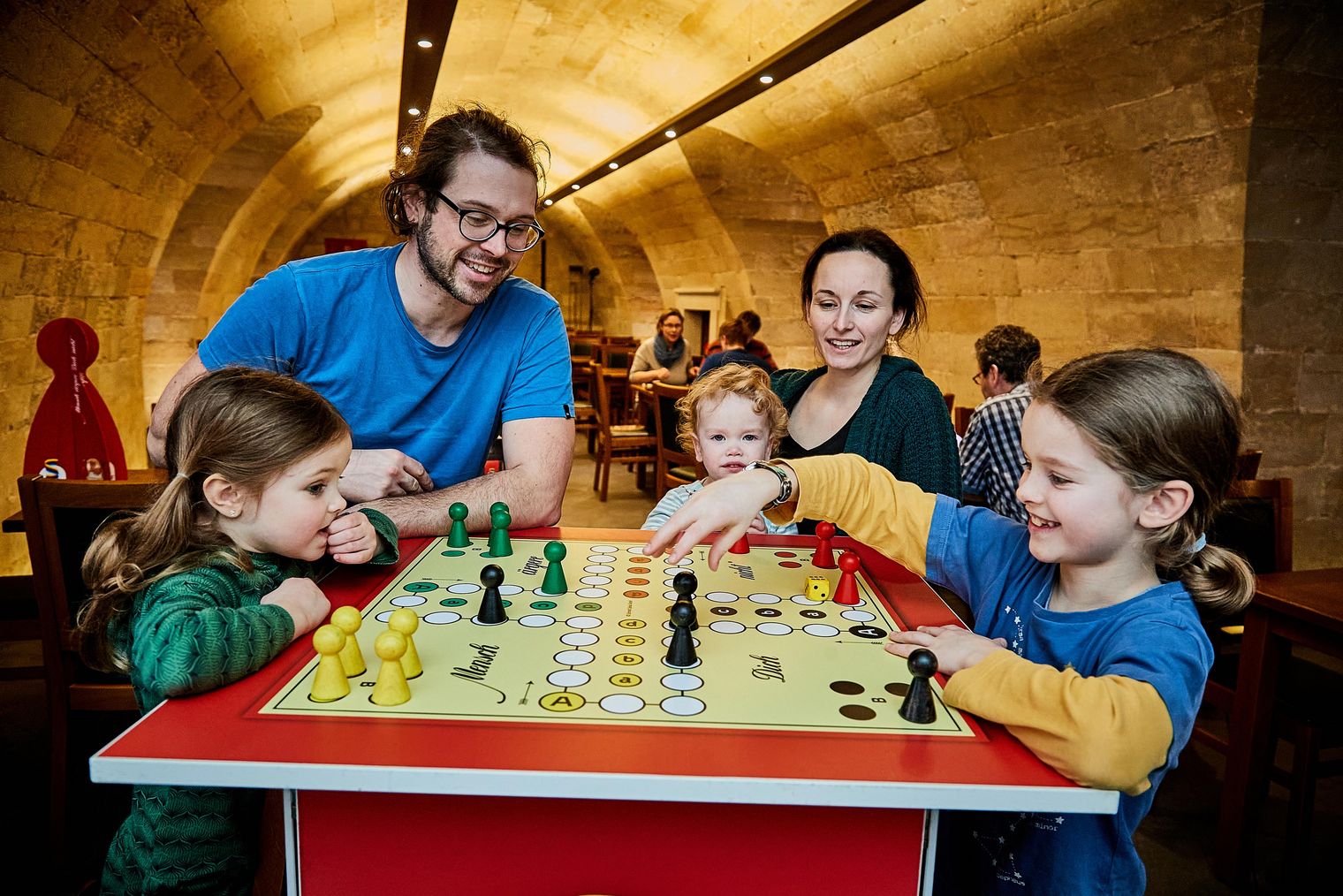Familie spielt gemeinsam an einem Tisch mit einem Brettspiel in einem gemütlichen Raum.