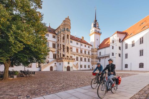 Zwei Radfahrer fahren mit Fahrrädern in Torgau mit dem historischen Gebäude des Schloss Hartenfels im Hintergrund.