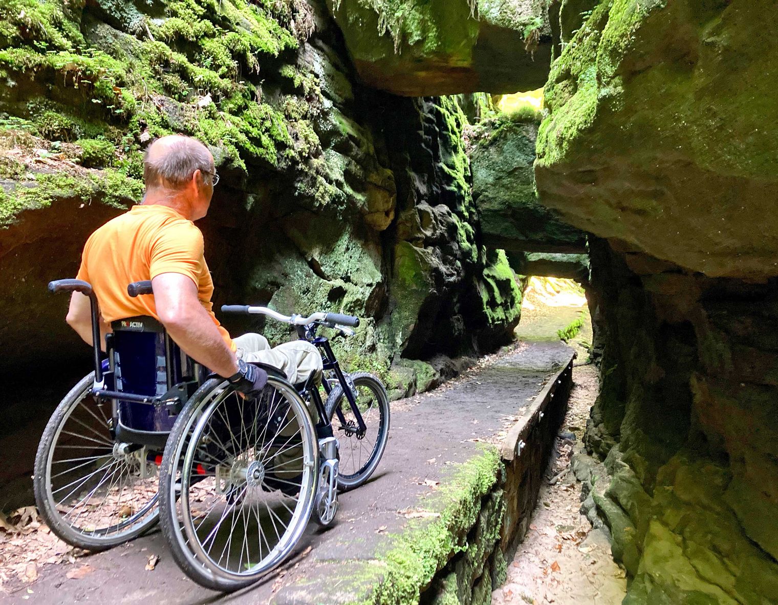 Mann im Rollstuhl auf einem schmalen Holzweg zwischen hohen, moosbedeckten Felsen.