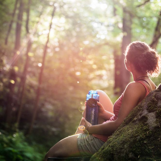 Frau sitzt auf einem Felsen im Wald, umgeben von Bäumen und sanftem Licht.