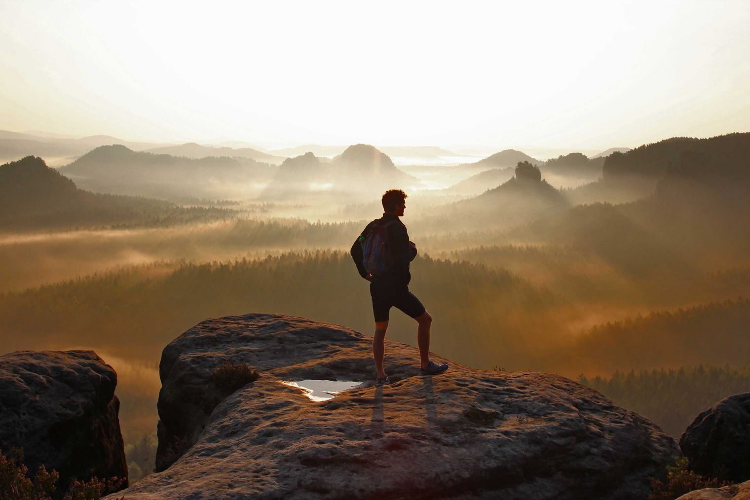 Person steht auf einem Felsen und blickt auf eine neblige Berglandschaft bei Sonnenaufgang.