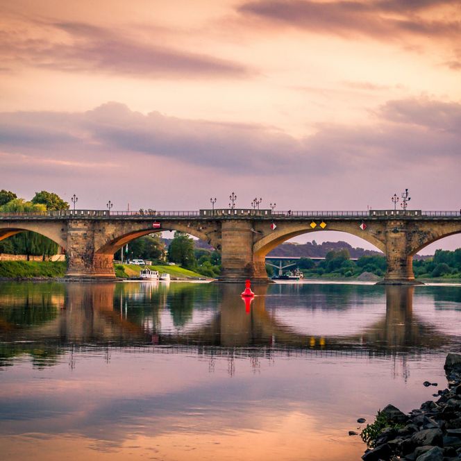 Brücke über einen ruhigen Fluss bei Sonnenuntergang, mit reflektierenden Farben im Wasser.