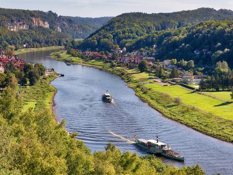 Flussufer von oben mit grünen Hügeln, Häusern und einem Dampfschiff unter klarem Himmel.