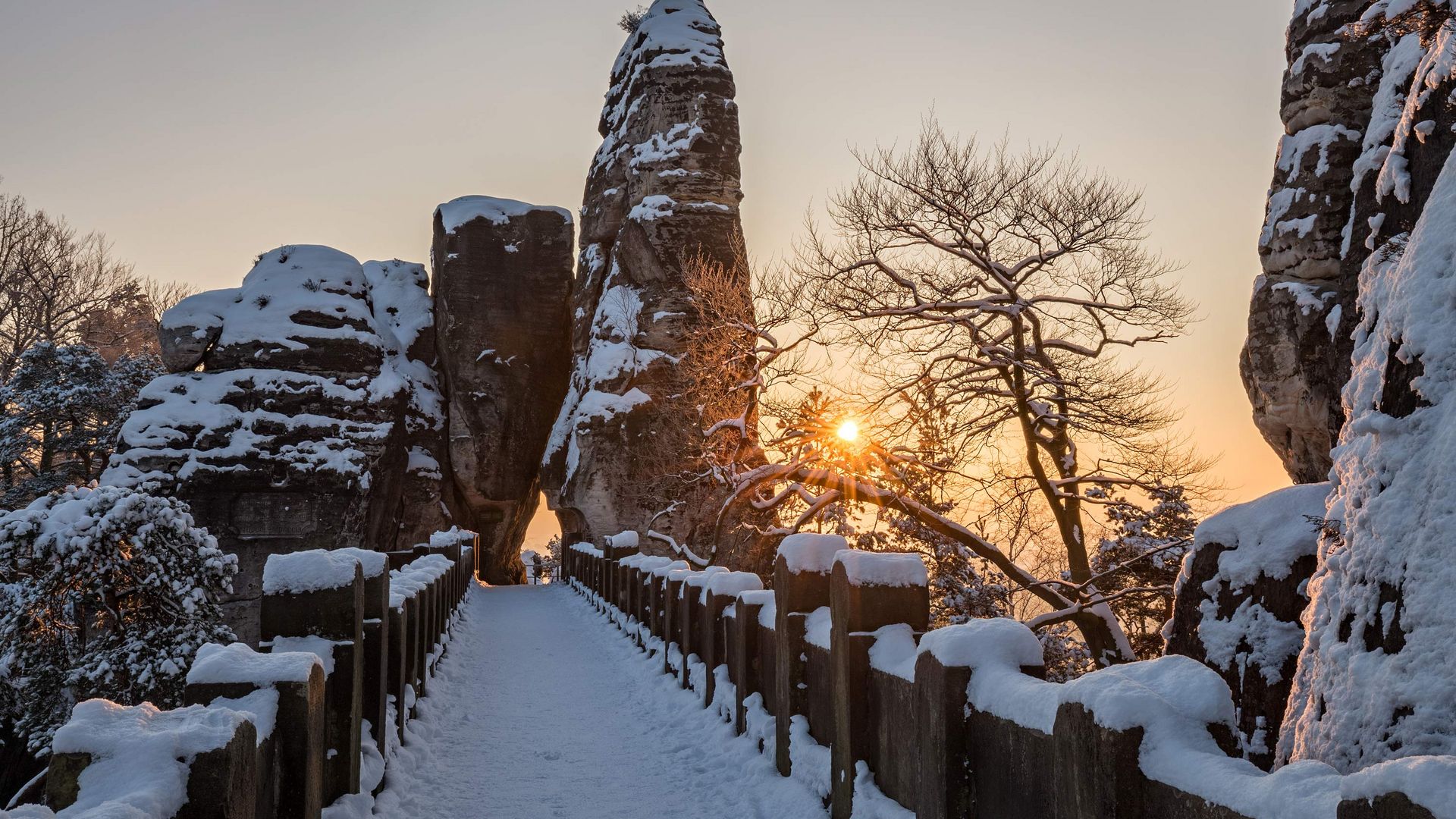Schneebedeckter Weg zwischen hohen Felsen, Sonnenaufgang im Hintergrund, Bäume im Vordergrund.