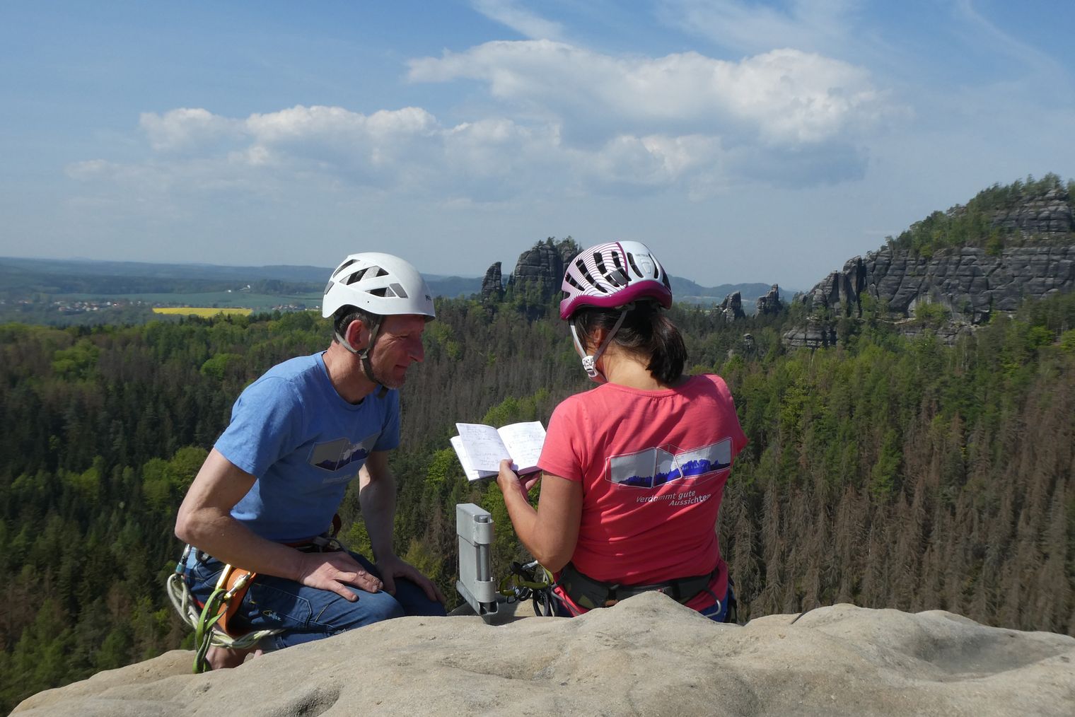 Zwei Kletterer mit Helmen betrachten eine Karte auf einem Felsen, Landschaft im Hintergrund.