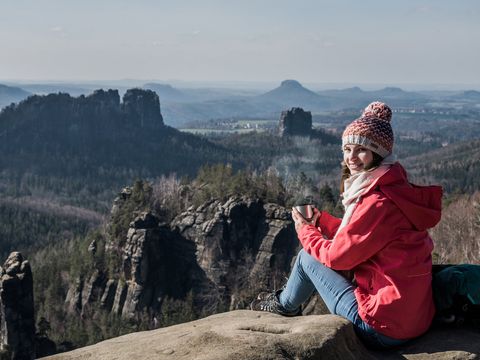 Frau mit Winterkleidung sitzt auf Carolafelsen und blickt auf bewaldete Sandsteinfelsen, blauer Himmel mit Wolken.