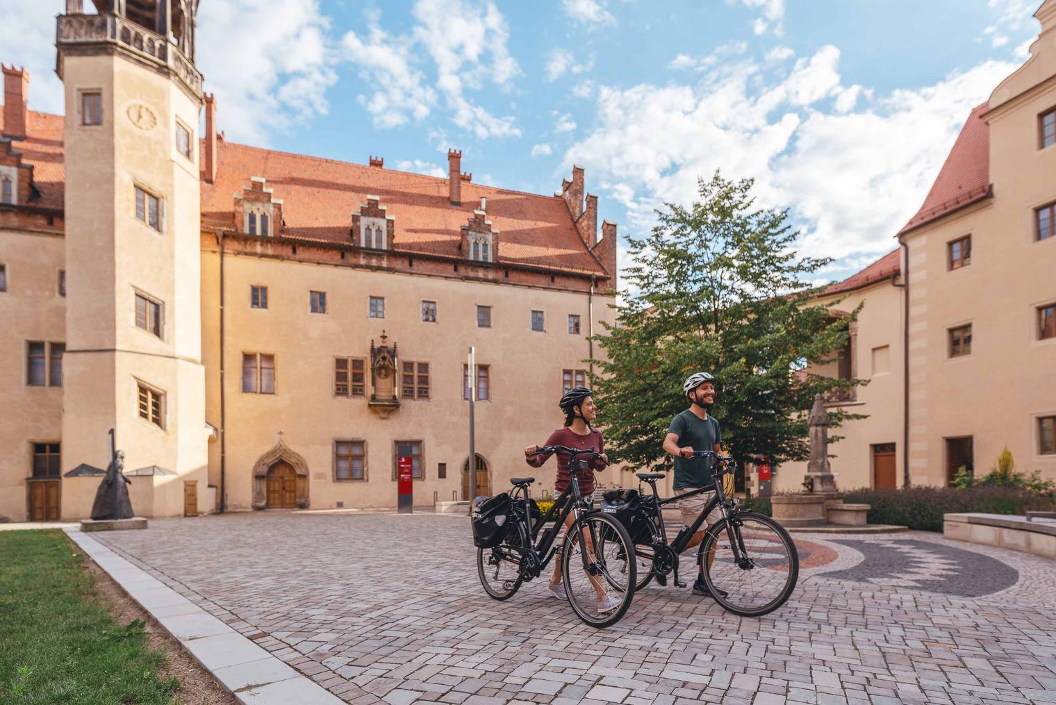 Historisches Gebäude mit orangefarbenem Mauerwerk, Bäumen und Fahrrädern im Vordergrund.