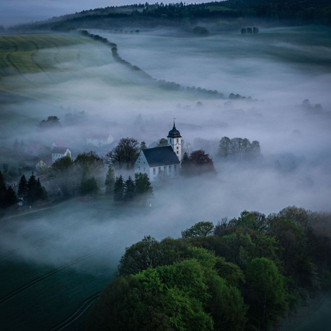 Eine Kirche steht umgeben von Nebel und Bäumen in einer hügeligen Landschaft.