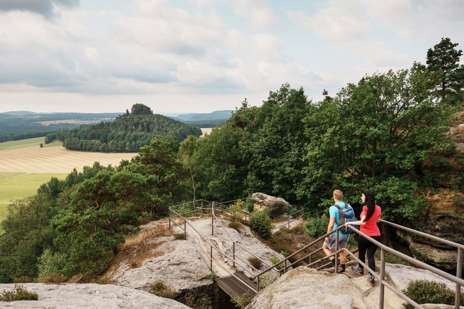 Zwei Wanderer auf einem Holzsteg mit Felsen, umgeben von Bäumen und einer weiten Landschaft.
