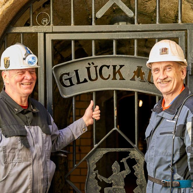 Zwei Männer in Schutzhelmen stehen vor einem Tor mit dem Schriftzug "GLÜCK".