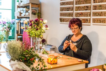 Frau mit Brille sitzt an einem Tisch und arrangiert Blumen, umgeben von Pflanzen und Werkzeugen.