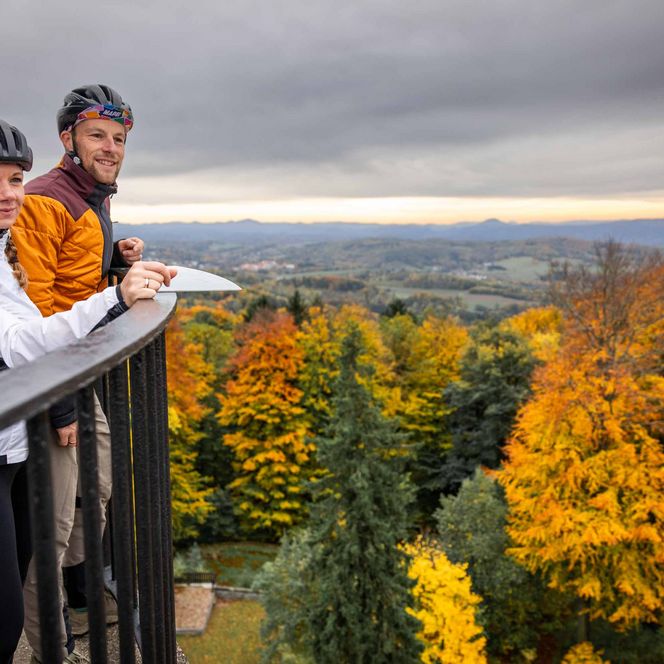 Paar mit Fahrrädern steht an einem Geländer und blickt auf eine herbstliche Landschaft mit bunten Bäumen.