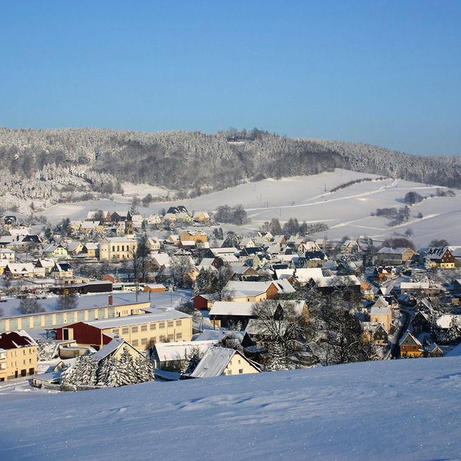 Schneebedeckte Landschaft mit einem Dorf und Bergen im Hintergrund unter klarem blauen Himmel.