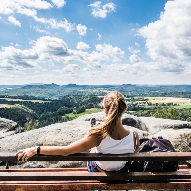 Frau sitzt auf einer Bank und blickt auf eine weite Landschaft mit Bergen und Wolken.