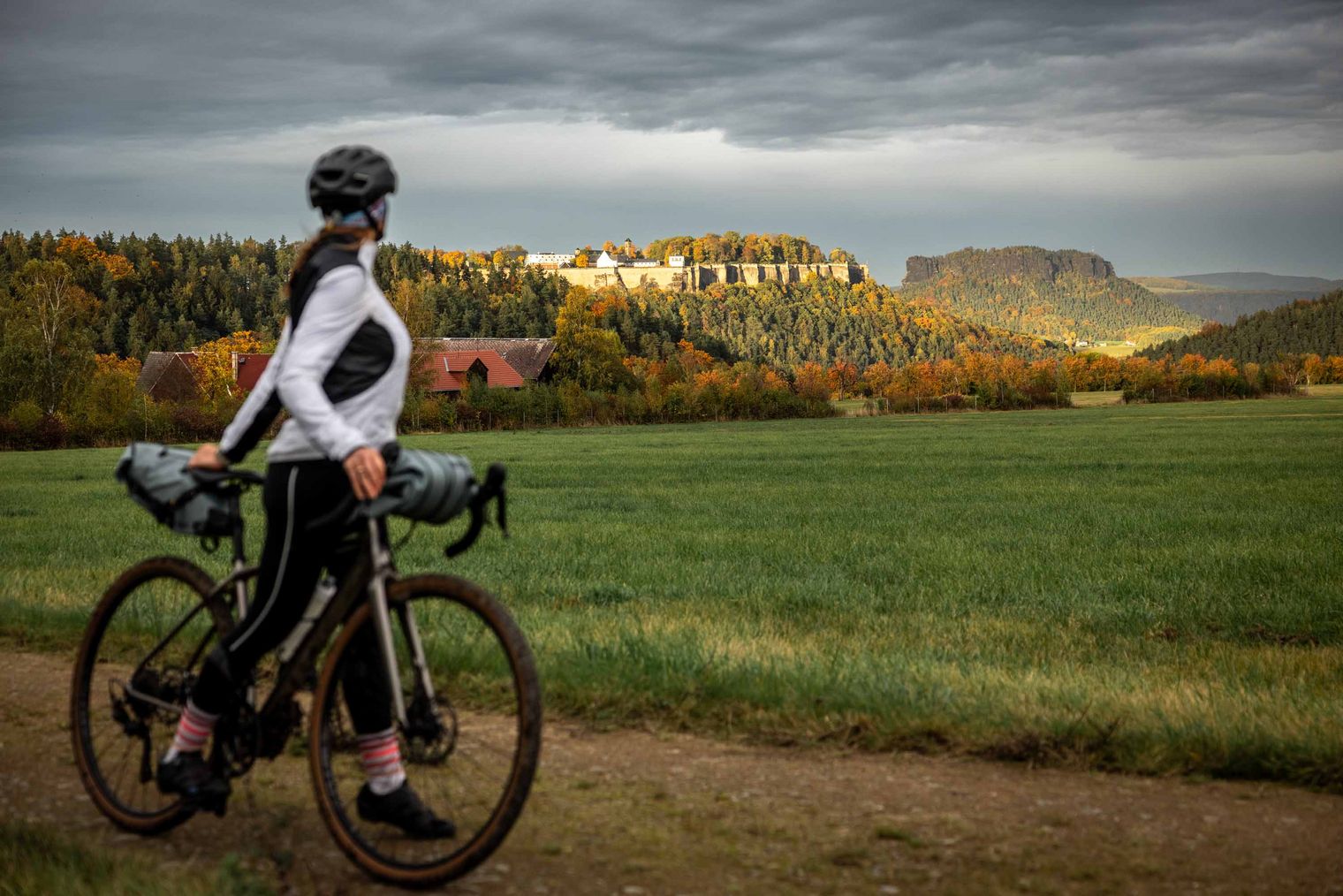 Radfahrerin in sportlicher Kleidung steht neben ihrem Fahrrad auf einem Feld, im Hintergrund der Tafelberg Lilienstein und die Festung Königstein und bewölkter Himmel.