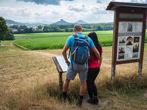 Zwei Wanderer stehen vor Infotafel auf Waldweg, im Hintergrund Wiesen und Wald.