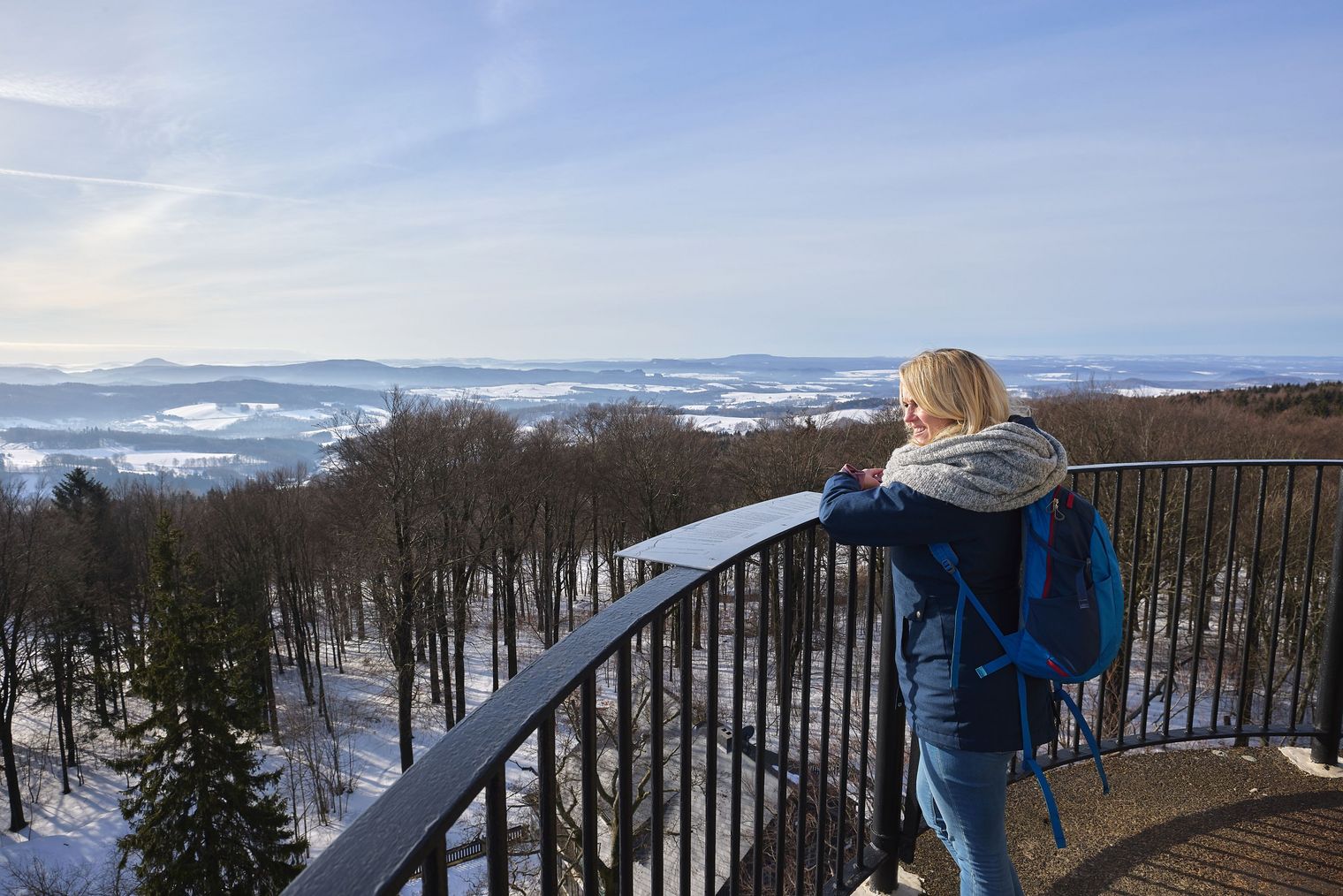 Person mit blonden Haaren steht an einem Geländer und blickt auf eine verschneite Landschaft.