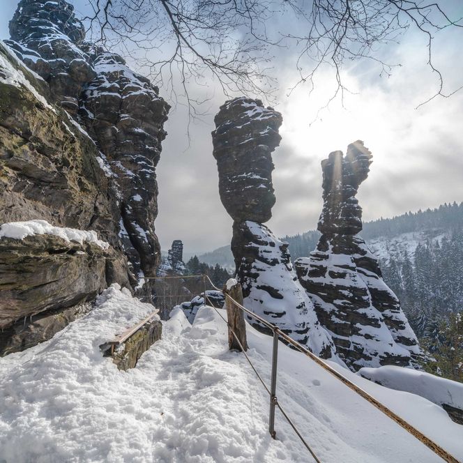 Schneebedeckte Felsenlandschaft im Bielatal mit Sonnenstrahlen und bewölktem Himmel im Hintergrund.