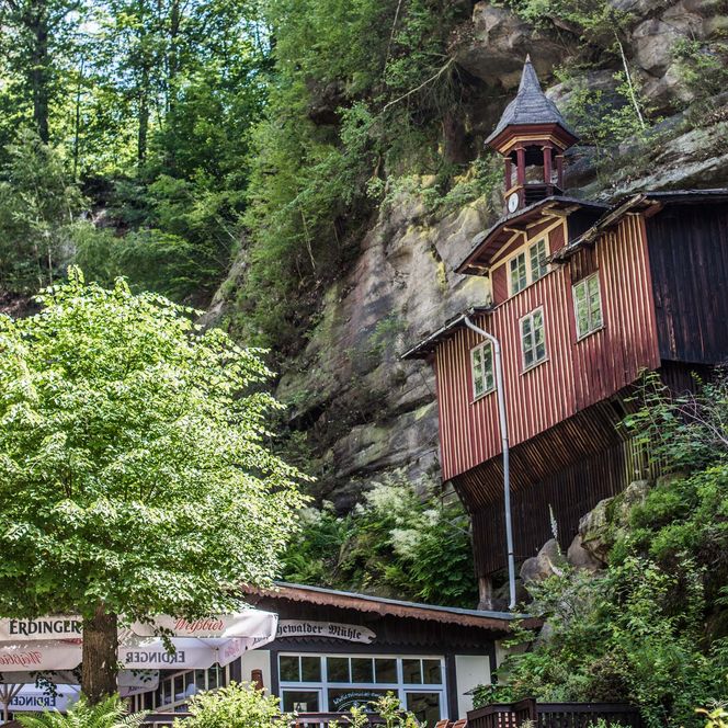 Holzhaus an einer Felswand, umgeben von Bäumen und einer Terrasse im Vordergrund.