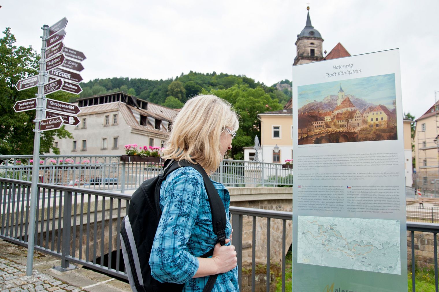 Frau mit Rucksack betrachtet eine Infotafel vor historischen Gebäuden und einer Kirche.