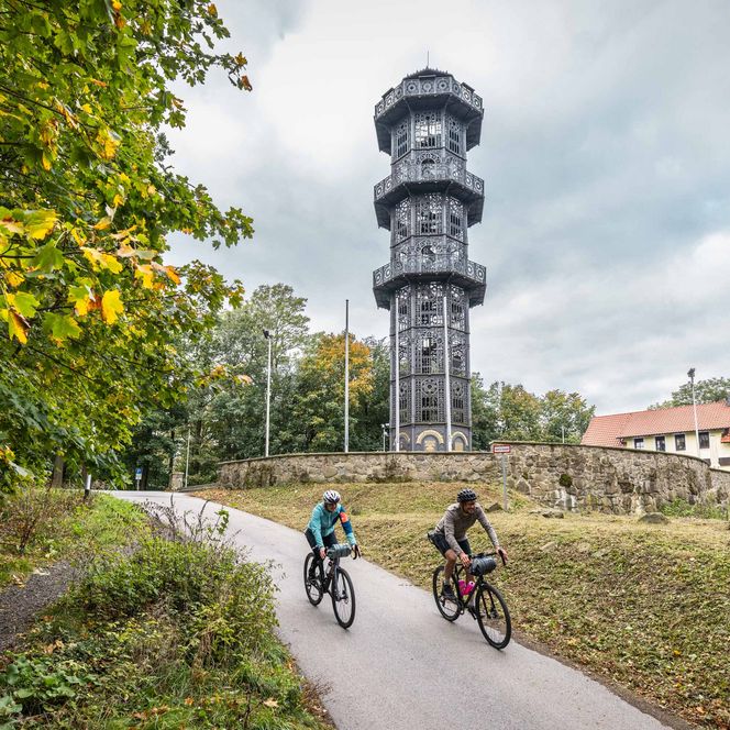 Radfahrer auf einem Weg auf dem Löbauer Berg neben einem hohen Turm, umgeben von Bäumen und einem bewölkten Himmel.