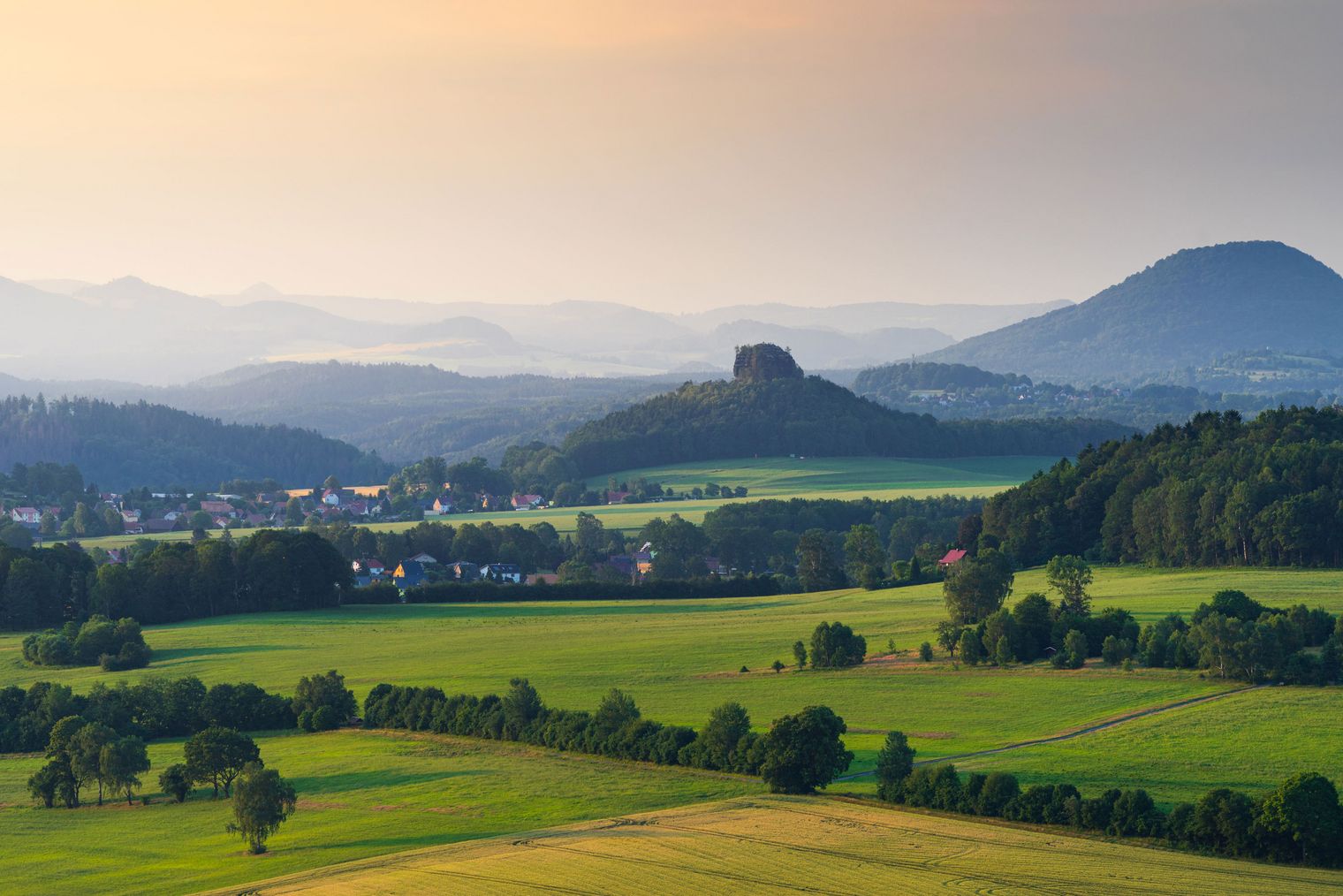 Blick auf eine hügelige Landschaft mit Feldern, Wäldern und einem markanten Felsen im Hintergrund.