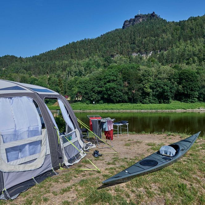 Zelt am Ufer eines Flusses, daneben ein Kajak und ein Grill, umgeben von grünen Bäumen und Hügeln.