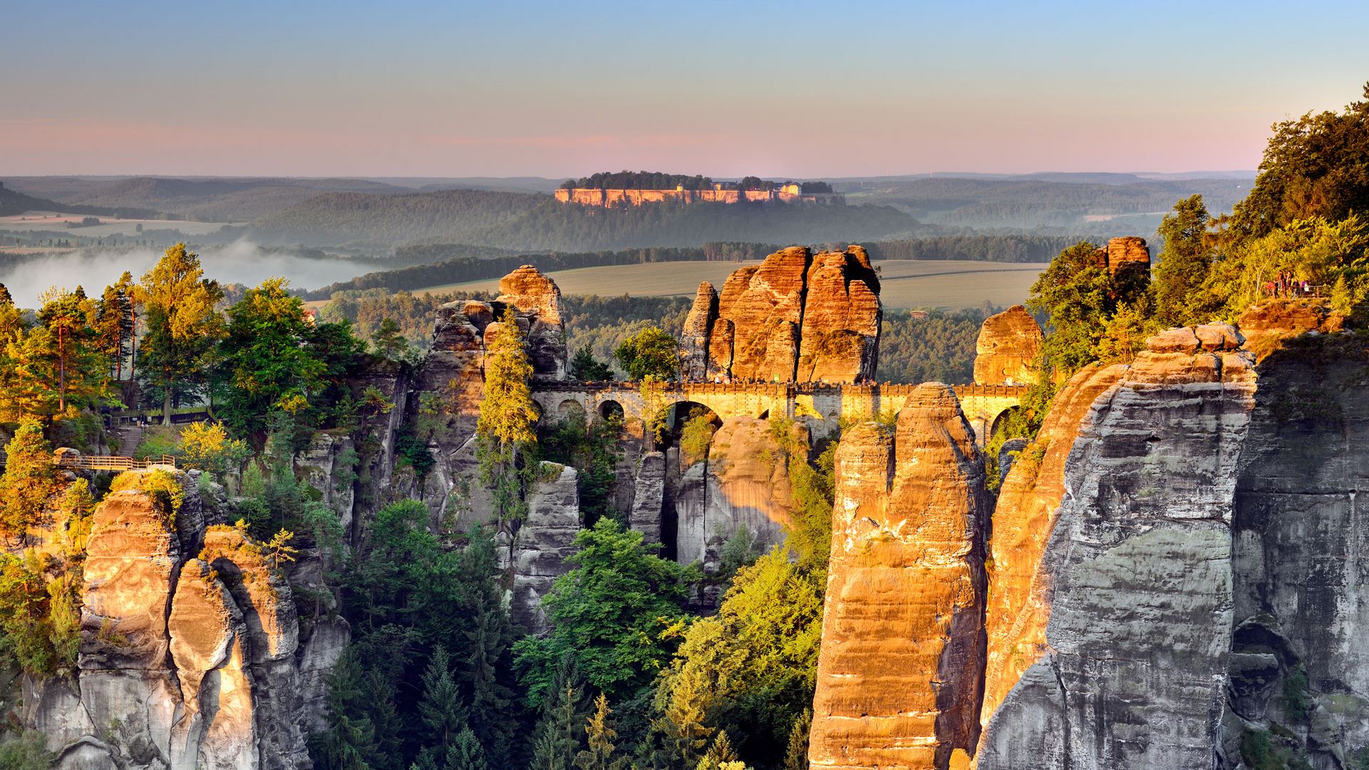 Felsenlandschaft Bastei mit Sonnenuntergang, bewaldete Hügel und ein weitläufiges Tal im Hintergrund.