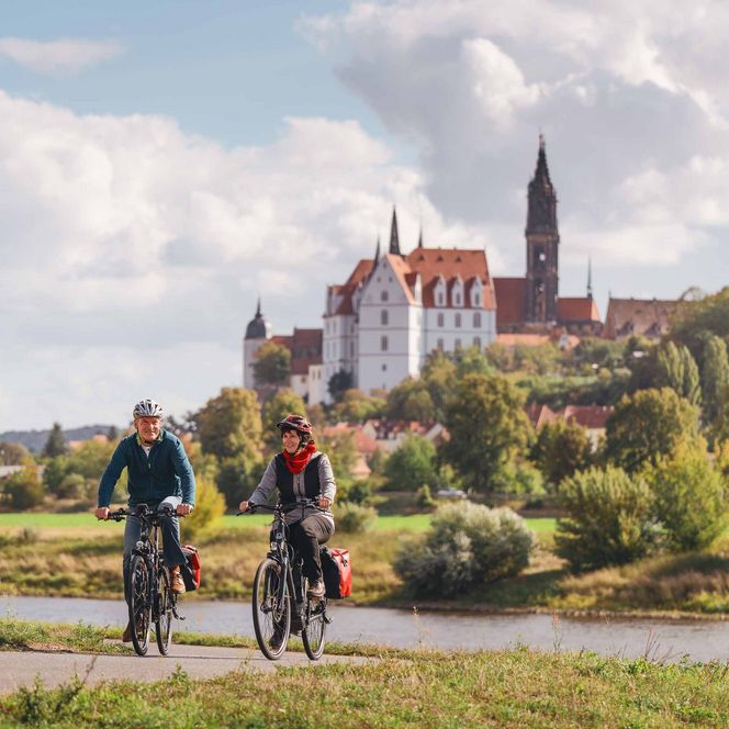 Zwei Radfahrer auf einem Weg mit einer Burg im Hintergrund und bewaldeten Hügeln.