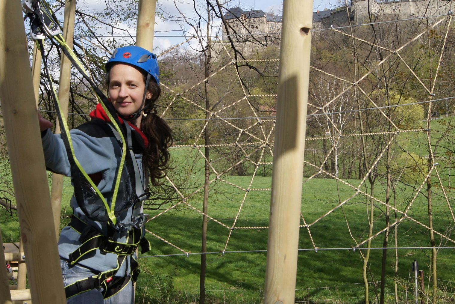 Person in blauem Helm hinter einem Netz im Kletterpark, lächelnd.