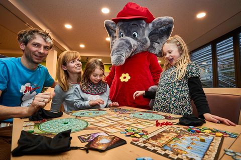 Familie spielt ein Brettspiel am Tisch, ein Elefantenmaskottchen steht daneben.