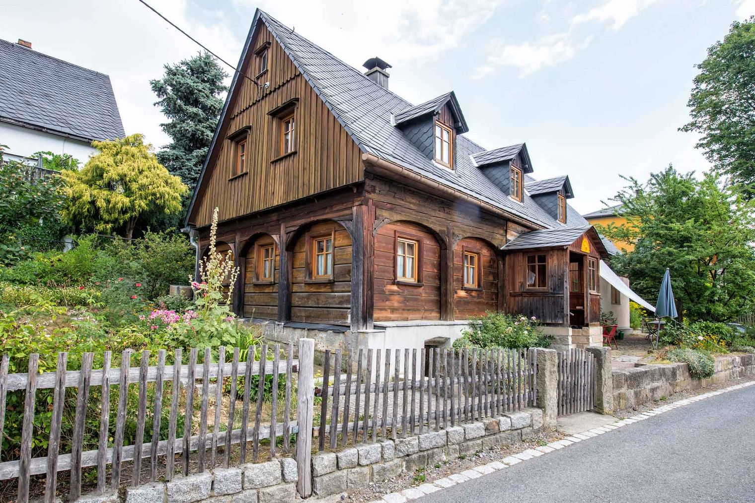 Holzhaus mit Satteldach, umgeben von Bäumen und einem Garten, eingezäunt mit einem Holzlattenzaun.
