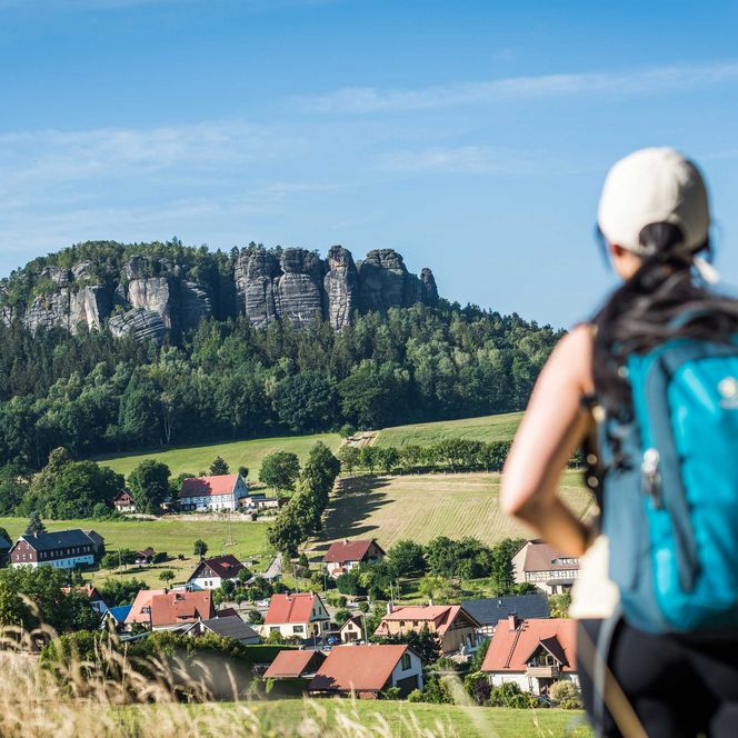 Wanderer mit Rucksack blickt auf den Pfaffenstein und eine grüne Landschaft mit Dörfern im Hintergrund.