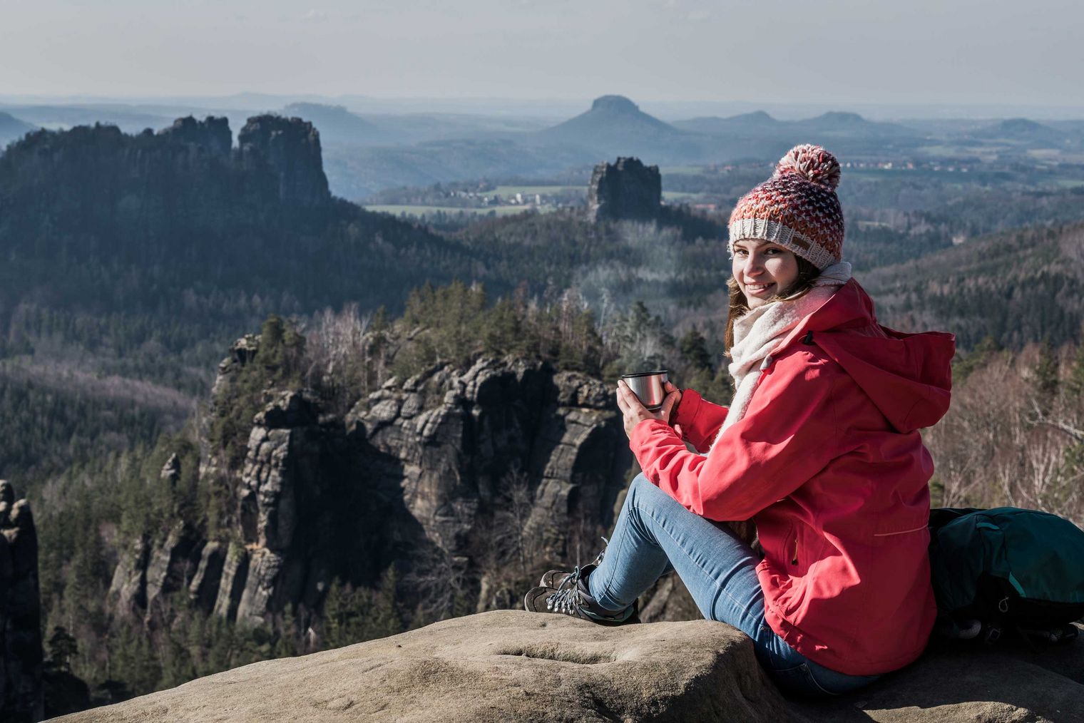 Frau in roter Jacke und Mütze sitzt auf einem Felsen mit Blick auf eine bergige Landschaft.