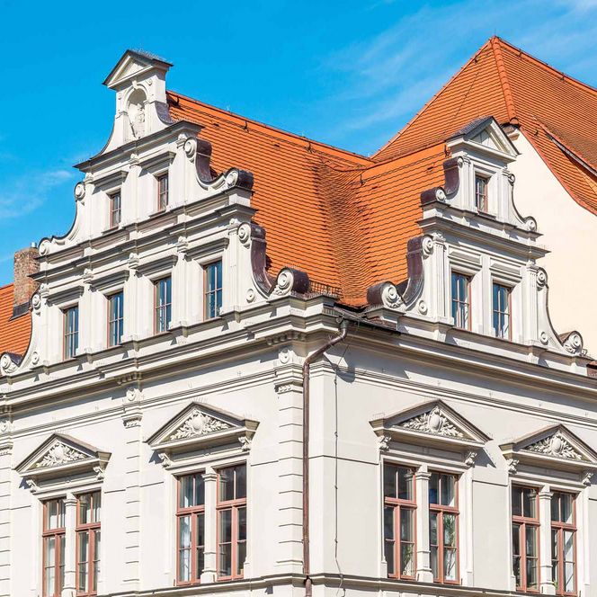 Historisches Gebäude mit weißer Fassade und rotem Satteldach unter blauem Himmel.
