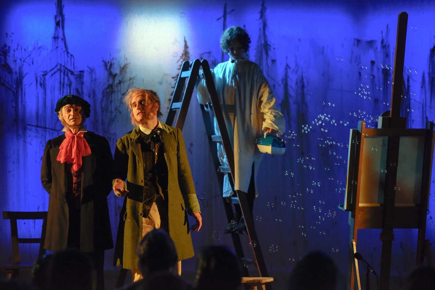 Szene aus Theateraufführung Richard Wagner Spiele: Zwei Schauspieler in Kostümen stehen auf einer Bühne, während ein dritter auf einer Leiter malt.