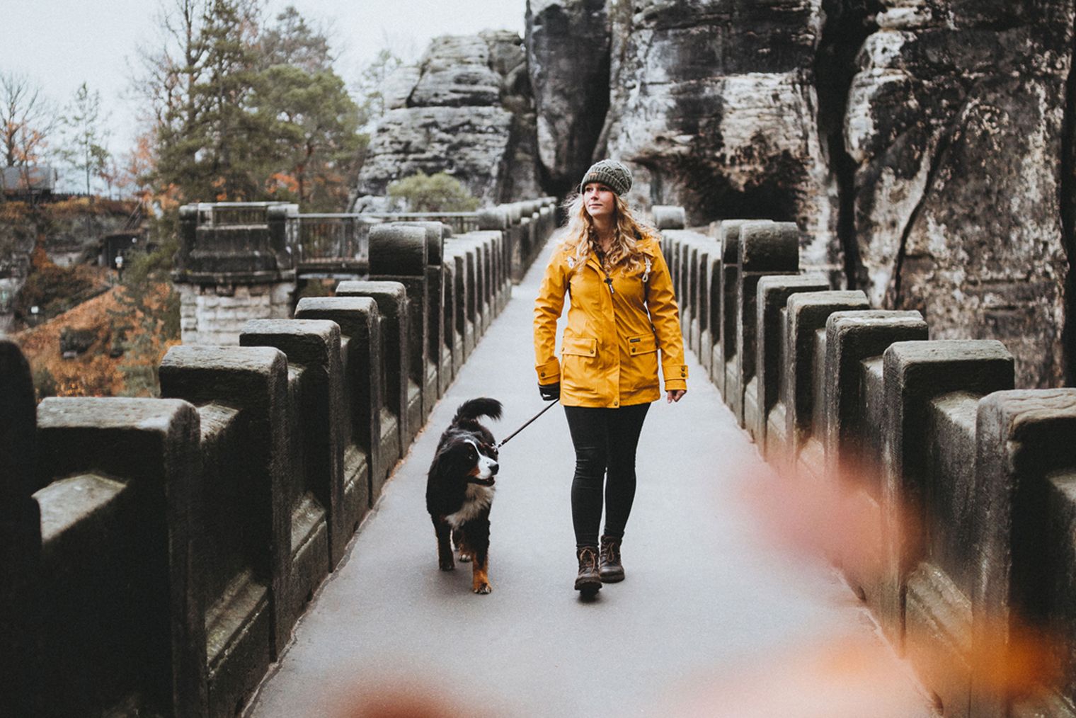 Frau in gelber Jacke geht mit Hund auf einer Brücke zwischen Felsen.