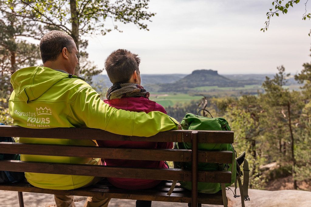 Paar sitzt auf einer Bank und blickt auf eine Landschaft mit Hügeln und Bäumen.