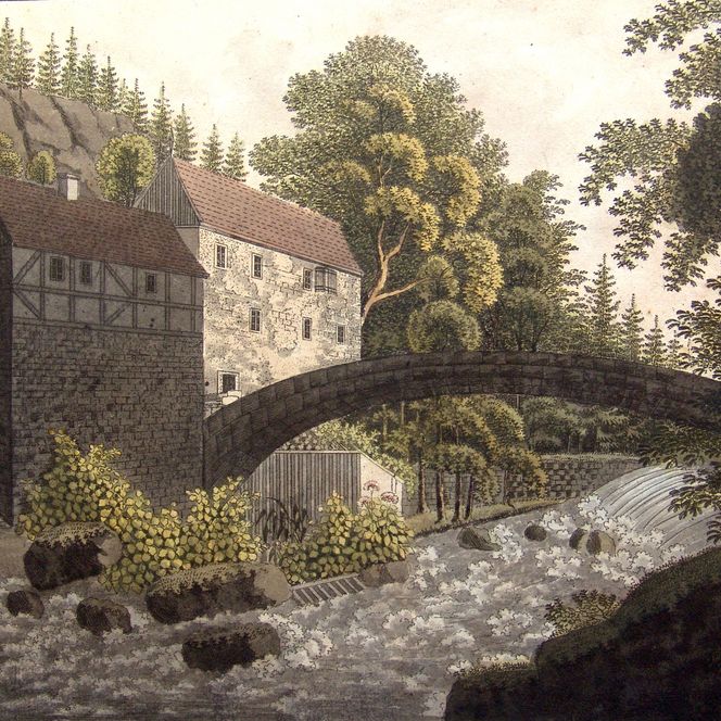 Historisches Gebäude neben einem Fluss, mit einer Steinbrücke und umgebenden Bäumen.