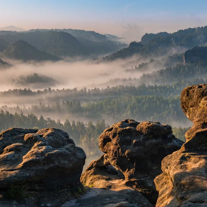 Blick auf eine neblige Landschaft mit Bergen und Wäldern im Hintergrund. Felsen im Vordergrund.