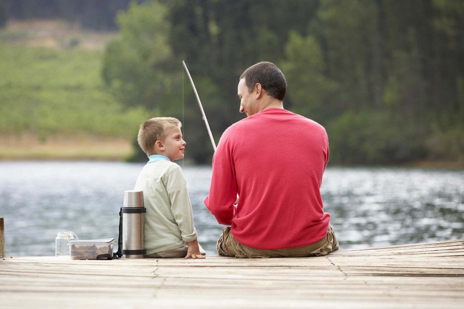 Vater und Sohn sitzen am Steg eines Sees und angeln, umgeben von Natur.