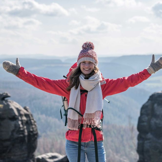 Frau in roter Jacke und Mütze steht mit ausgebreiteten Armen auf einem Felsen, Blick auf die Landschaft.