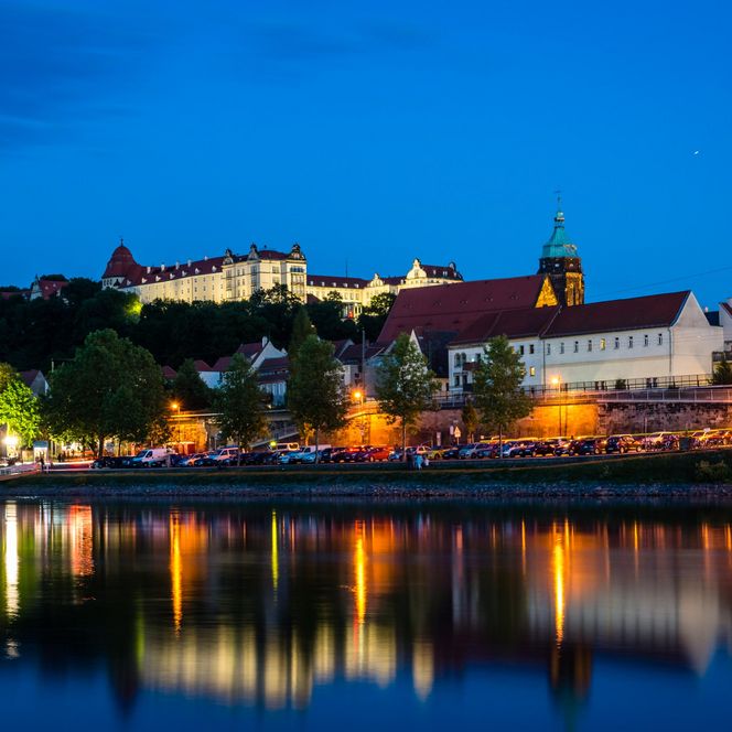 Blick auf eine Stadt am Wasser bei Nacht, mit beleuchteten Gebäuden und einer Burg im Hintergrund.