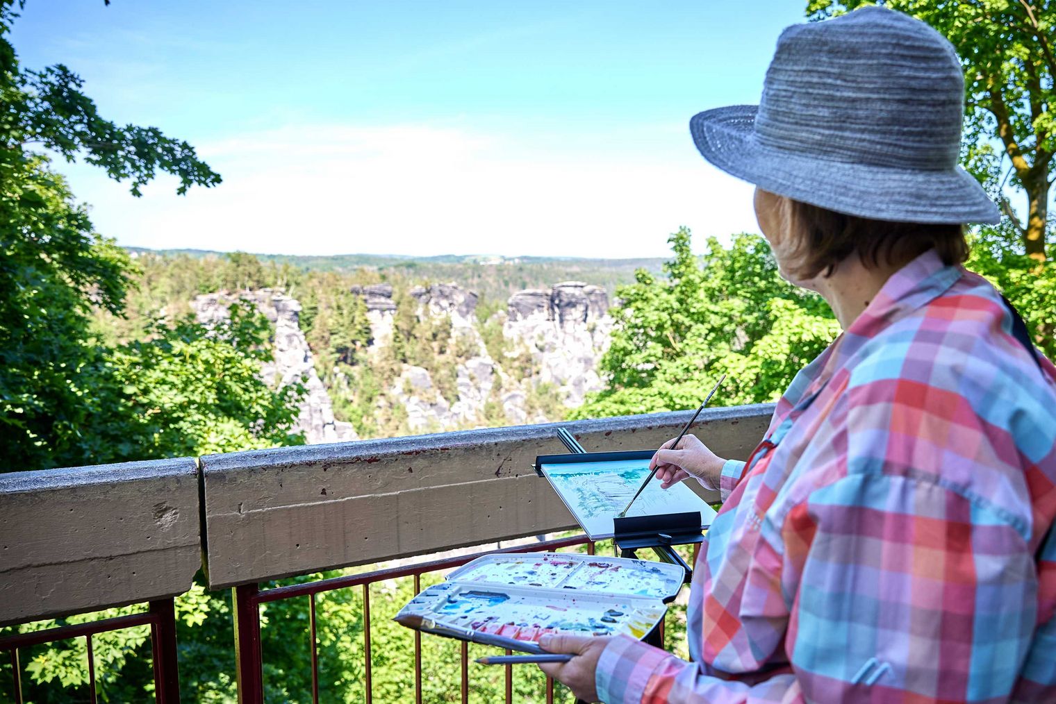 Frau mit Hut malt Landschaft von einer Aussichtsplattform aus, umgeben von Bäumen.