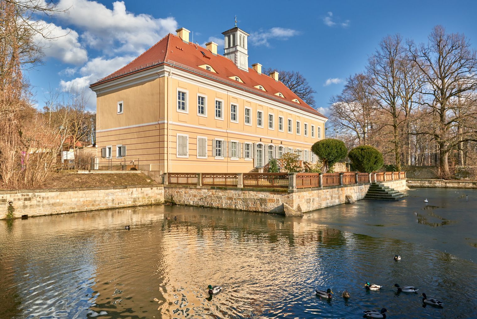 Historisches Gebäude mit rotem Dach, umgeben von Wasser und Bäumen, im Hintergrund blauer Himmel.